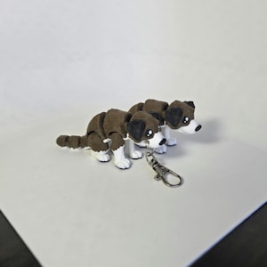 May include: Two brown and white dog figurines with black ears and noses are attached to a silver keychain. The dogs are positioned side-by-side, appearing to be looking down. The background is a plain white surface.