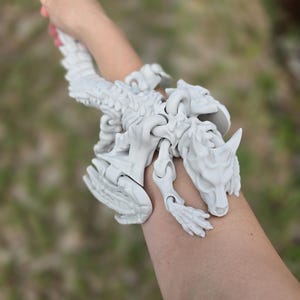 May include: A light gray, articulated dragon wrist cuff. The dragon has a detailed, skeletal design with a head, legs, and a long tail. The cuff is worn on a person's arm, with a blurred green background.