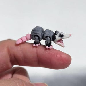 May include: A miniature articulated opossum figurine featuring a grey body, a white face with black eye markings, a pink nose, and a segmented pink tail. The small creature has an open mouth and pink feet, showcasing a detailed 3D printed design.