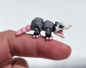 May include: A miniature articulated opossum figurine featuring a grey body, a white face with black eye markings, a pink nose, and a segmented pink tail. The small creature has an open mouth and pink feet, showcasing a detailed 3D printed design.