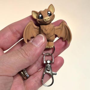 May include: A brown bat-shaped keychain with a silver clasp. The bat has large ears, wings, and a smiling face with big eyes. The keychain is a cute, whimsical accessory.