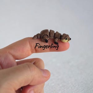 Könnte beinhalten: Eine kleine, bewegliche Otter-Figur in Braun und Beige mit segmentiertem Körper und schwarzen Augen. Das Wort Fingerling ist in schwarzer Schreibschrift unter der Figur aufgedruckt. Der Otter hat ein hellbeiges Gesicht und einen dunkelbraunen Körper.