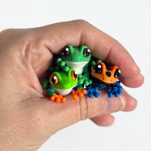 May include: Three colorful frog figurines. One green frog with orange feet and large eyes, another green frog with white eyes, and a third frog with an orange head, blue feet, and black eyes. The frogs are held in a hand.