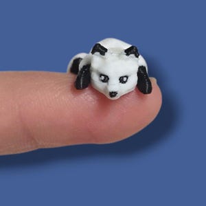 May include: A miniature white fox figurine with black ears and paws rests on a fingertip against a blue background. The small, detailed fox has a relaxed pose, with its head slightly raised. The figurine is a small decorative item.