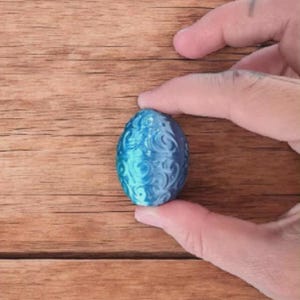 May include: A blue decorative egg with a swirling pattern. The egg is held between two fingers against a wooden background. The egg's color is a gradient of light and dark blue.