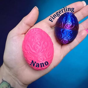 May include: Two small egg-shaped containers with intricate embossed swirl patterns. One egg is a matte hot pink with the word Nano printed in black, and the other is a metallic blue and purple gradient with the word Fingerling printed in black.