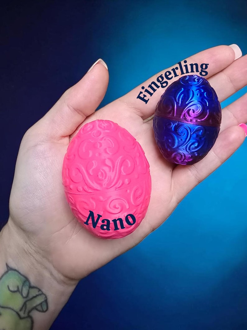 May include: Two small egg-shaped containers with intricate embossed swirl patterns. One egg is a matte hot pink with the word Nano printed in black, and the other is a metallic blue and purple gradient with the word Fingerling printed in black.