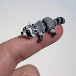 May include: A miniature articulated raccoon figurine featuring a gray and white body with black eye patches and a striped tail. The small, detailed creature has a segmented design that allows for movement, showcasing a realistic appearance.