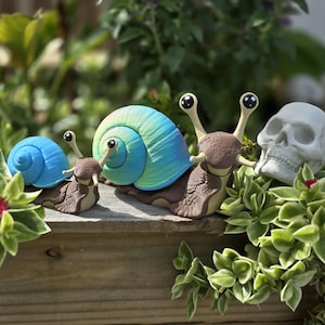 May include: A whimsical garden scene features three snail figurines with colorful shells. Two snails have blue and teal shells, while the third is a skull. The snails are brown and sit among green plants on a wooden surface.