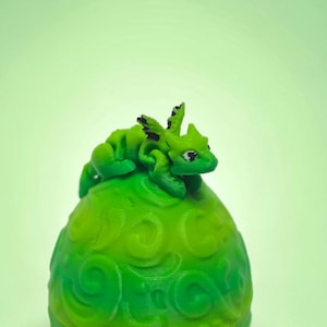 May include: A small, bright green articulated dragon figurine with black wing accents perched atop a larger, rounded green egg-shaped object featuring a raised, swirling embossed pattern across its entire surface.