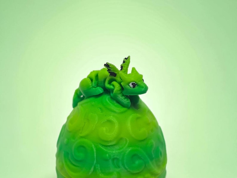May include: A small, bright green articulated dragon figurine with black wing accents perched atop a larger, rounded green egg-shaped object featuring a raised, swirling embossed pattern across its entire surface.