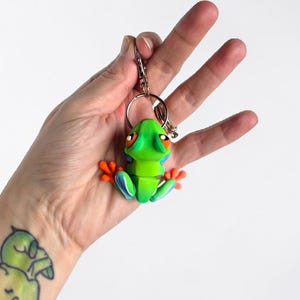 May include: A green frog keychain with orange feet and large orange eyes. The frog has blue accents and is attached to a silver metal keyring and clasp. The frog is held against a white background.