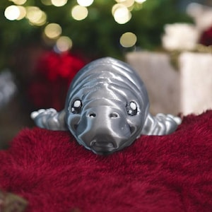 May include: A silver-colored manatee figurine with a textured surface, resting on a plush, deep red surface. The manatee has small, detailed eyes and flipper-like appendages. The background features blurred Christmas lights and greenery.
