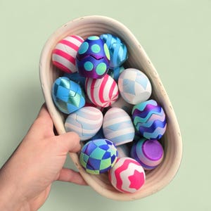 May include: A hand holding a woven basket filled with colorful, patterned Easter eggs. The eggs feature designs in shades of pink, blue, purple, and white. The basket has a light brown color, and the background is a soft green.