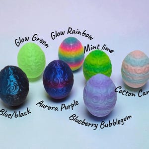 May include: Seven egg-shaped objects with embossed swirl patterns. Colors include Glow Green, Glow Rainbow, Mint Lime, Cotton Candy, Blue/Black, Aurora Purple, and Blueberry Bubblegum. Each egg is labeled with its corresponding color name in black text.