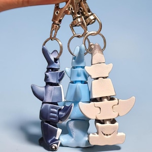 May include: Three keychains with a totem-like design in shades of blue and white. Each keychain features a series of interlocking shapes, with a silver clasp at the top. The keychains are held against a light blue background.