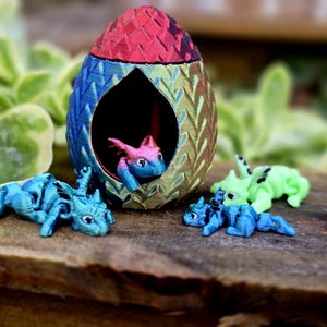 May include: A colorful dragon egg with a red top, blue and gold sides, and a dragon inside. Several small blue and green dragons are scattered around the egg on a wooden surface. The scene is set outdoors.