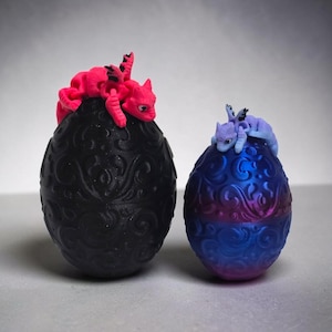 May include: Two decorative eggs with intricate embossed scrollwork patterns. A matte black egg is topped with a bright pink articulated dragon figure, while a smaller metallic blue and purple egg features a light purple articulated dragon figure.