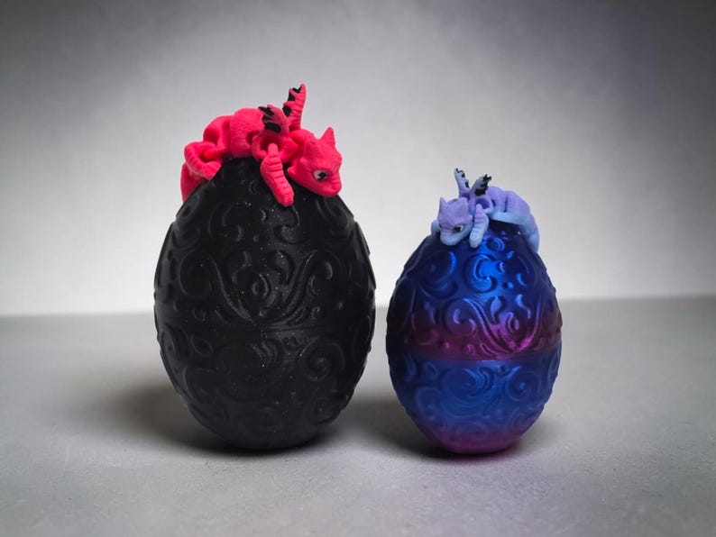 May include: Two decorative eggs with intricate embossed scrollwork patterns. A matte black egg is topped with a bright pink articulated dragon figure, while a smaller metallic blue and purple egg features a light purple articulated dragon figure.