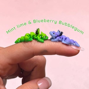 May include: Two small articulated dragon figurines, one lime green and one periwinkle blue, each featuring black dorsal spikes and detailed facial features. The text Mint lime & Blueberry Bubblegum is arched above the two miniature creatures.