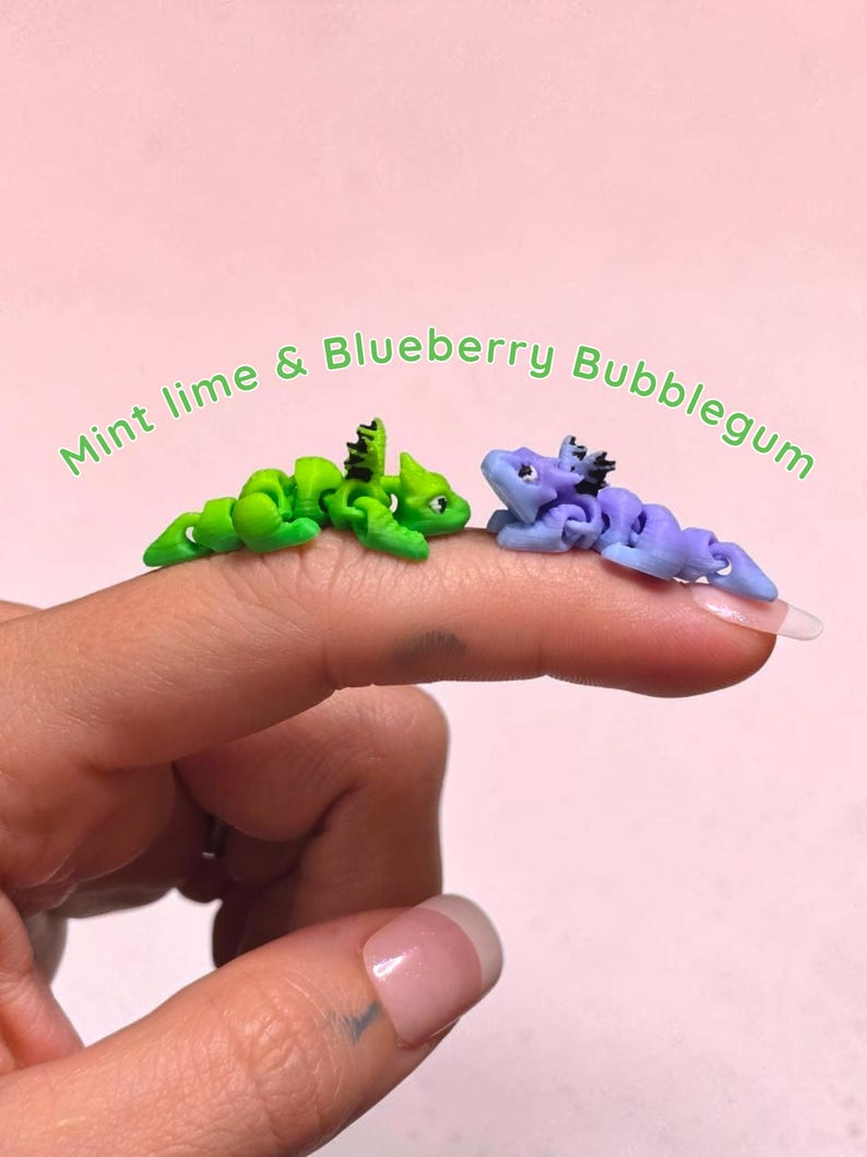 May include: Two small articulated dragon figurines, one lime green and one periwinkle blue, each featuring black dorsal spikes and detailed facial features. The text Mint lime & Blueberry Bubblegum is arched above the two miniature creatures.