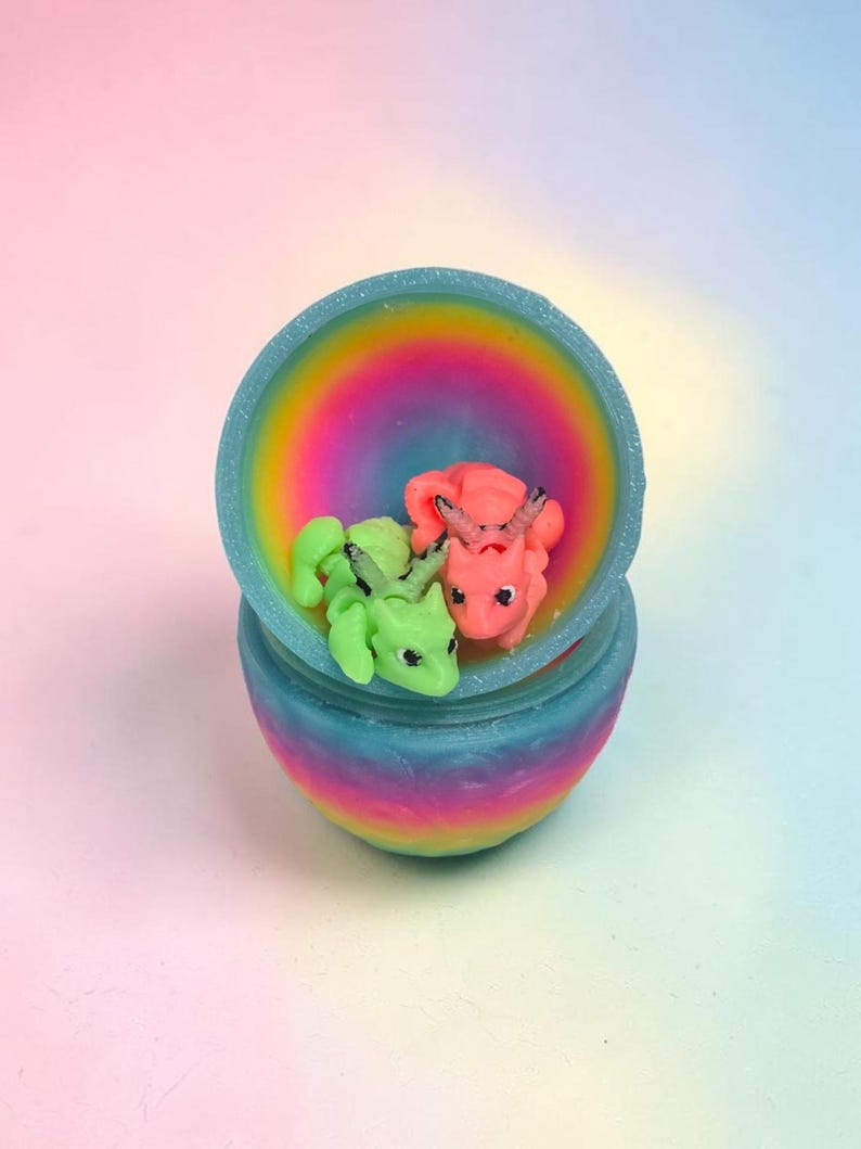 May include: A small, round, rainbow-colored container with an open lid, holding two miniature dragon figurines. One dragon is bright lime green with dark accents, and the other is a vibrant coral pink with dark horns and facial details.