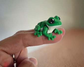 May include: A small, green frog figurine with large black eyes and white accents. The frog is perched on a finger, showcasing its detailed features and vibrant colors. The figurine's design highlights its small size and playful nature.