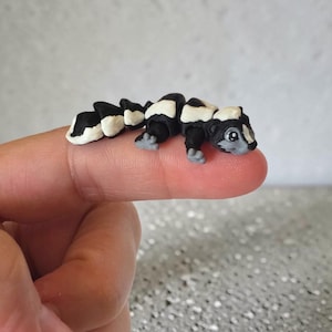 May include: A miniature sculpted figurine of a skunk with a black and white patterned body, a segmented tail, and small grey feet. The creature has a grey face with a white stripe, black ears, and large dark eyes, crafted in a small scale.