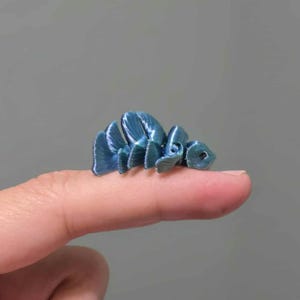 Könnte beinhalten: Eine kleine, bewegliche blaue Fischfigur, die auf einer Fingerspitze ruht. Der Fisch hat einen segmentierten Körper und Flossen mit metallischem Glanz. Der Hintergrund ist einfarbig grau.