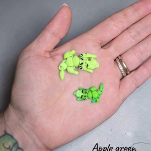 May include: Two small, articulated, 3D-printed raccoon figures in apple green and mint lime colors. Each figure features a segmented body, a detailed head with black painted eye markings, and a curled tail. The text Apple green & mint lime is visible.