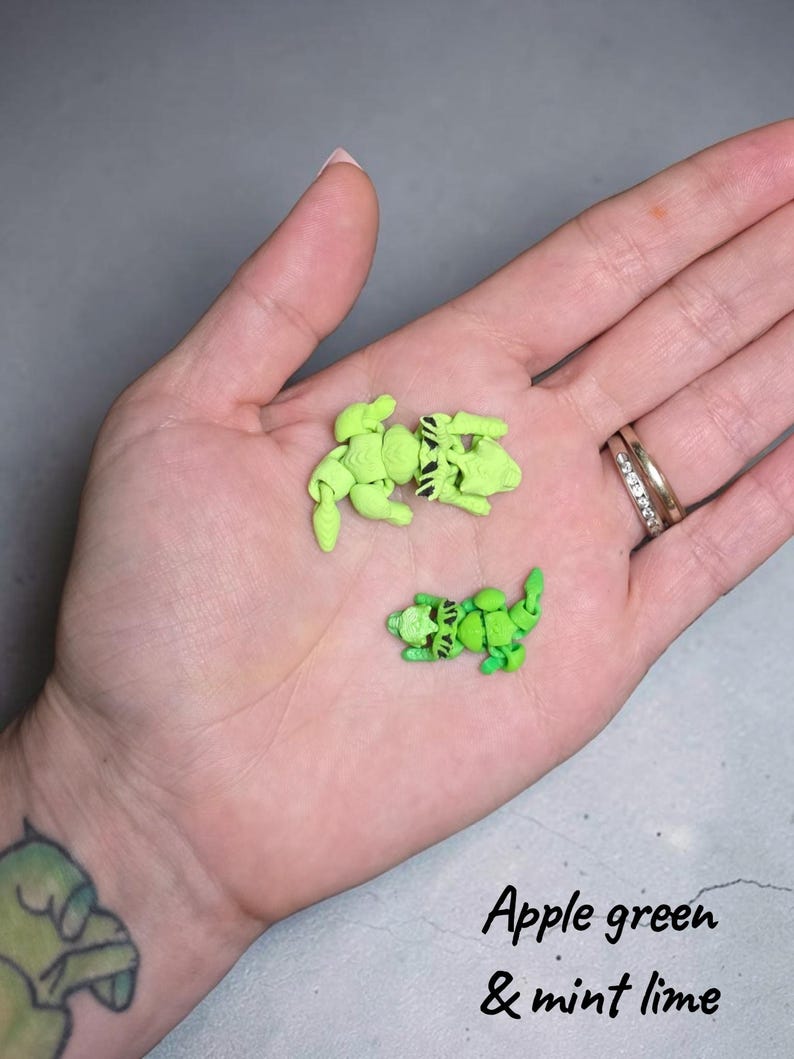 May include: Two small, articulated, 3D-printed raccoon figures in apple green and mint lime colors. Each figure features a segmented body, a detailed head with black painted eye markings, and a curled tail. The text Apple green & mint lime is visible.