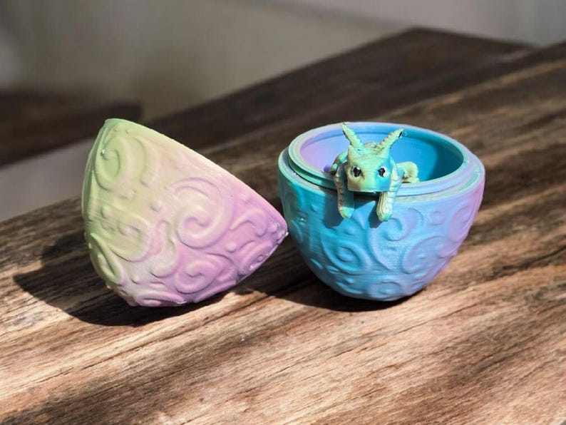 May include: A two-piece spherical container with a pastel gradient of yellow, pink, and blue featuring a raised swirling pattern. A small, light green creature with pointed ears and dark eyes is perched on the rim of the open bottom section.