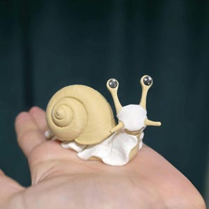 May include: A detailed figurine of a snail with a cream-colored shell and a white body. The snail has two long, slender antennae with small, round eyes. The figurine is resting on a surface.
