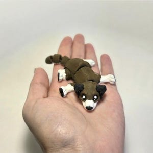 Pode incluir: Uma pequena figura de cão feita à mão em castanho, preto e branco. O cão está deitado com as patas abertas. A figura está na palma de uma mão.