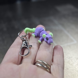 May include: A handmade keychain featuring a small, purple creature with green accents. The creature has a whimsical design, with a long tail and small feet. The keychain includes a silver-colored clasp and ring for attaching to keys or bags.