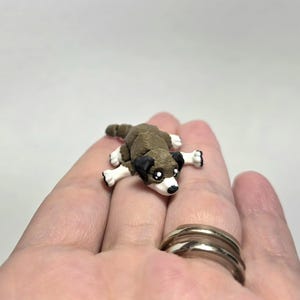 Pode incluir: Uma figura em miniatura de um cão, deitado com as patas estendidas. O cão é castanho e branco com detalhes pretos nas orelhas e ao redor dos olhos. A figura está em uma mão.