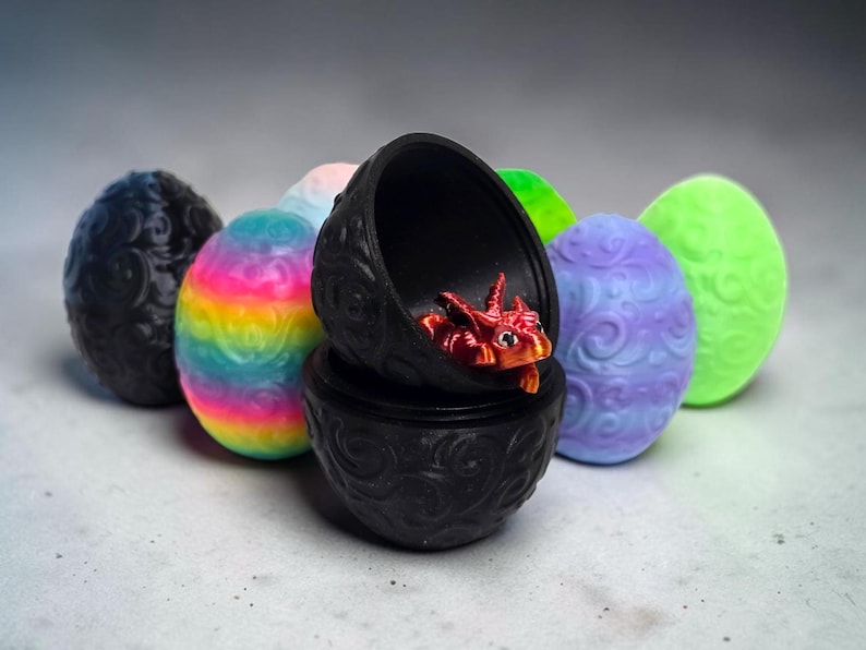 May include: A collection of oval-shaped containers with embossed swirl patterns in various colors, including black, rainbow, purple, and neon green. One black container is open, revealing a small, metallic red dragon figurine inside.
