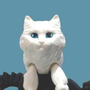Op de afbeelding: Een witte kattenfiguur met helderblauwe ogen en een pluizig uiterlijk. De kat staat tegen een blauwgroene achtergrond. Het beeldje heeft een gedetailleerd, getextureerd oppervlak.