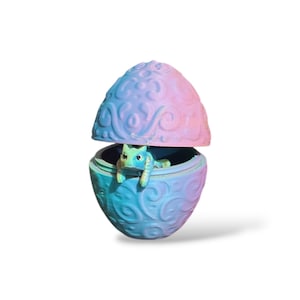 May include: A small, two-piece egg-shaped container featuring an embossed swirling pattern in a gradient of pink, purple, and blue. A small, light green creature with visible eyes and limbs is positioned inside the bottom half of the egg.