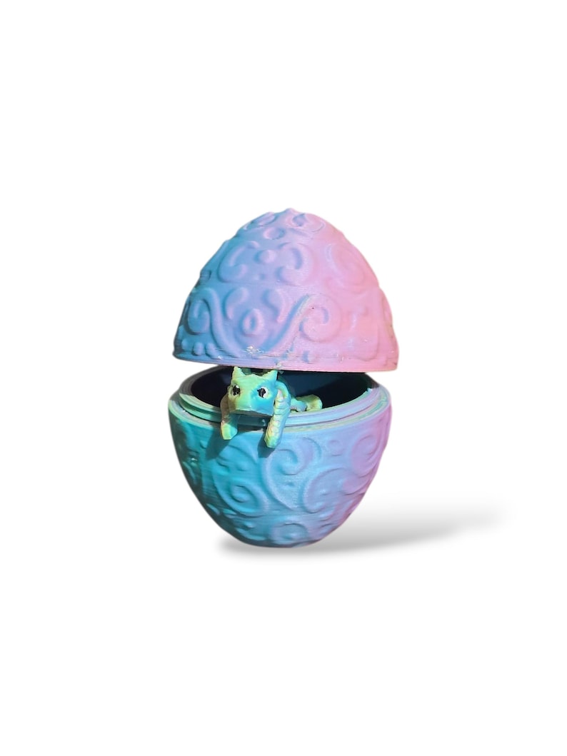May include: A small, two-piece egg-shaped container featuring an embossed swirling pattern in a gradient of pink, purple, and blue. A small, light green creature with visible eyes and limbs is positioned inside the bottom half of the egg.