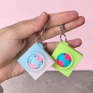 Könnte beinhalten: Zwei bunte Schlüsselanhänger. Einer ist hellblau mit einem rosa und blauen Design, der andere ist limettengrün mit einem rosa und blauen Design. Beide haben einen silbernen Verschluss und eine quadratische Form.