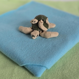 May include: A small, handcrafted turtle figurine made of clay. The turtle has a dark green shell and tan-colored head, flippers, and tail. It rests on a light blue fabric, with a green fabric in the background. The turtle is a decorative item.