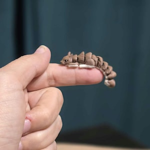 May include: A small, articulated brown squirrel figurine with a segmented body and a fluffy tail. The miniature sculpture is resting on a finger, showcasing its detailed design and craftsmanship. The squirrel has a light brown color with white accents.