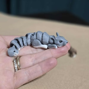 May include: A grey articulated squirrel figurine with a white belly and black eyes. The toy is made of segmented pieces, allowing for movement. The squirrel is resting on a hand.