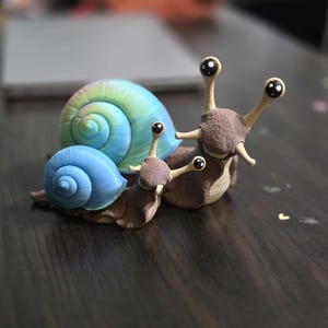 Pode incluir: Uma família de estatuetas de caracóis. O maior caracol tem uma concha azul e verde em espiral, enquanto os caracóis menores têm conchas azuis. Os corpos dos caracóis são castanhos com antenas longas e finas e olhos grandes e expressivos.