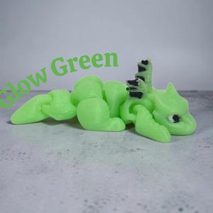 May include: A 3D printed articulated dragon figurine in a light glow green color with black accents on the eyes and dorsal spines. The text Glow Green is printed in green letters above the dragon, which features a segmented body design.