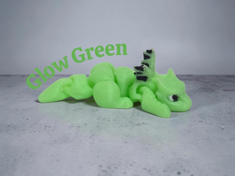 May include: A 3D printed articulated dragon figurine in a light glow green color with black accents on the eyes and dorsal spines. The text Glow Green is printed in green letters above the dragon, which features a segmented body design.