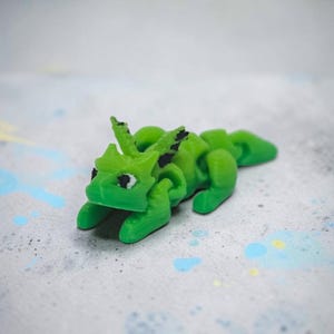 May include: A small, articulated dragon figurine in a vibrant lime green color. The creature features a segmented body, pointed ears, and black painted eyes. The surface of the dragon shows fine horizontal lines consistent with 3D printing.
