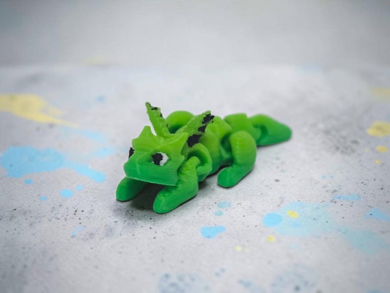 May include: A small, articulated dragon figurine in a vibrant lime green color. The creature features a segmented body, pointed ears, and black painted eyes. The surface of the dragon shows fine horizontal lines consistent with 3D printing.