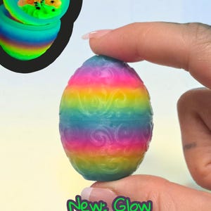 May include: A rainbow-colored egg with a raised swirl pattern. The egg is shown closed and also open in an inset, revealing a small, multi-colored creature inside. The text "New: Glow Rainbow" is printed in purple at the bottom of the image.